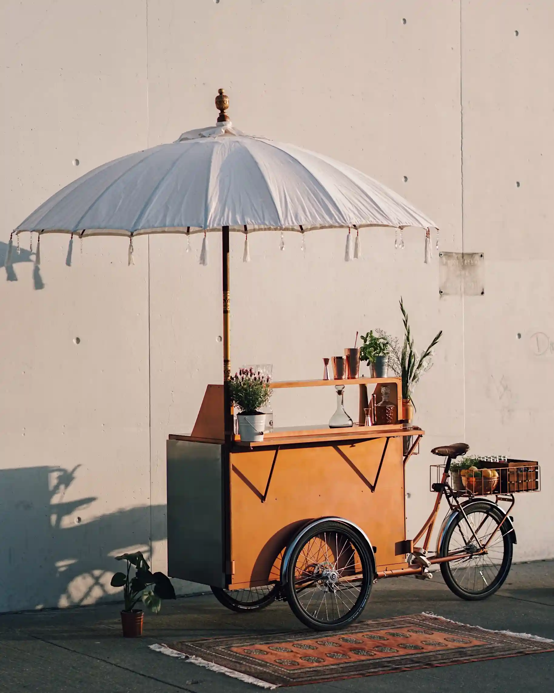 Eine kupferfarbene mobile Fahrrad-Bar auf drei Rädern mit großem, weißen Schirm.